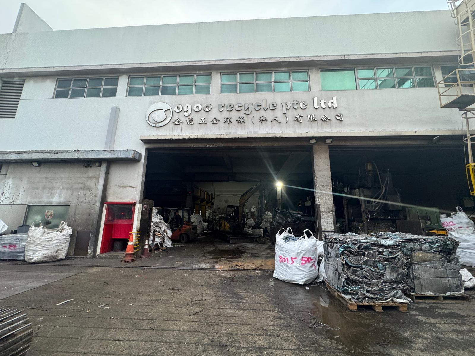 The Ogoo Recycle Pte Ltd yard at Tuas View Close, Singapore — covered processing hall with excavator, forklift and baling press, and baled metal stacked outside